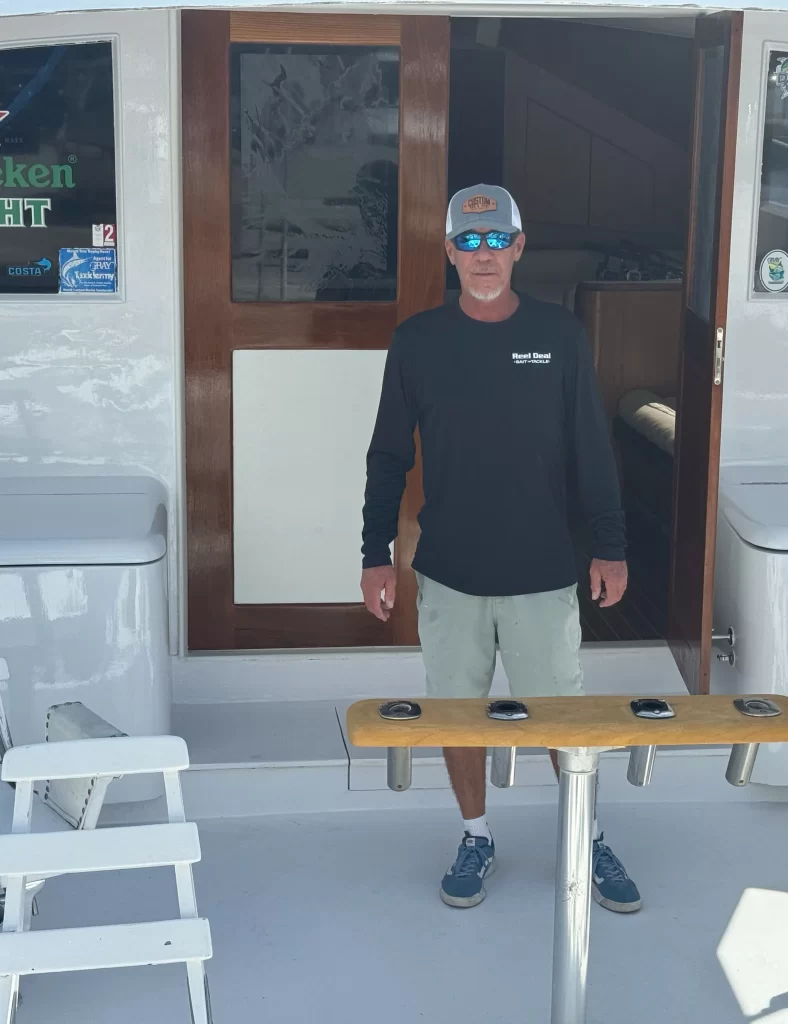 A man in a dark long-sleeve shirt, light shorts, baseball cap, and sunglasses stands on a boat deck, ready for pompano drift fishing. There's a wooden door and seating area behind him. The boat's interior is partially visible through the open door as they set off from Pompano Beach.