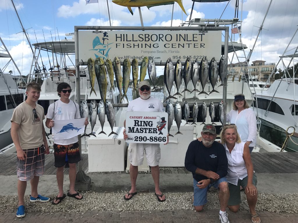 A group of people is posing with freshly caught fish at the Hillsboro Inlet Fishing Center, renowned for its pompano drift fishing. The fish are displayed on a rack behind them, and a sign with contact details reads 