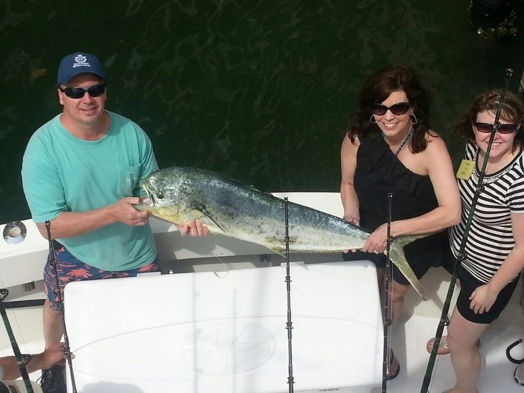 Three people on a boat proudly display a large fish. The man on the left wears a blue cap and green shirt, while the two women are dressed in black. The woman on the right has sunglasses. The scene suggests a successful fishing trip in sunny weather.