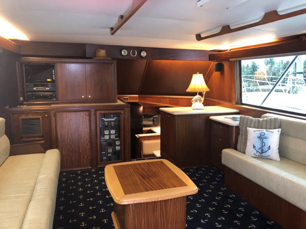 The image showcases the yacht's cabin with wooden furniture, a lamp on the cabinet, and a cushioned seating area adorned with an anchor-themed pillow. The carpet features anchor prints, while windows offer a view of Hillsboro Inlet fishing charters at the marina.