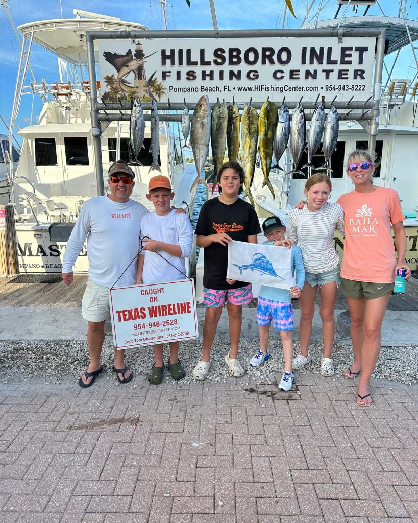 A group of six people stand smiling in front of a fishing charter sign near Hillsboro Inlet. They're holding up freshly caught fish and a sign that reads 