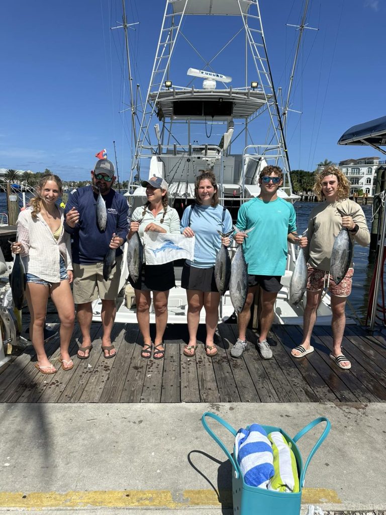 A group of six people stands on a dock in front of a boat, each proudly holding a large catch from their Pompano Beach adventure. They celebrate the successful drift fishing trip under the clear, sunny sky, while a blue basket with towels rests on the ground nearby.