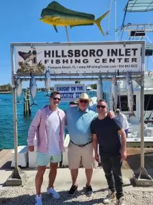 Three people pose in front of a fishing sign at Hillsboro Inlet Fishing Center, showcasing their catch from a day of pompano drift fishing. They stand under several fish hanging from the rack, with scenic water views and a docked boat in the background.