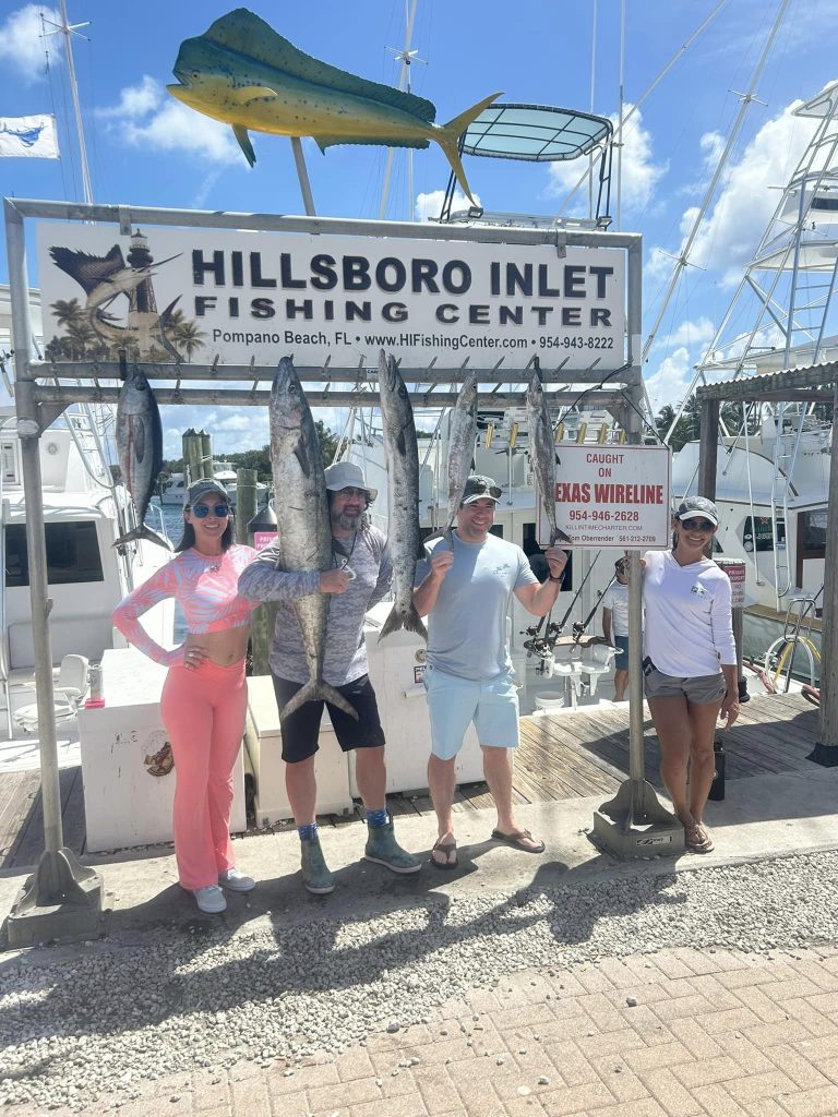 Four people stand smiling with large fish hanging beside them at Hillsboro Inlet Fishing Center, well-known for Hillsboro Inlet fishing. A boat and a sign with contact information are visible in the background. The sky is clear with a few clouds, perfect for pompano drift fishing.