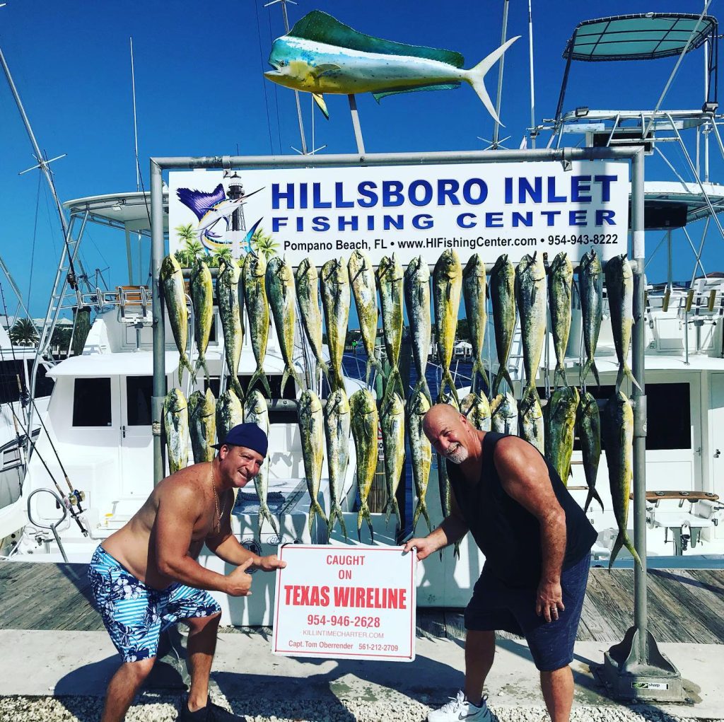 Two men pose at Hillsboro Inlet Fishing Center, surrounded by a display of impressive fish caught while pompano drift fishing. They hold a sign reading 