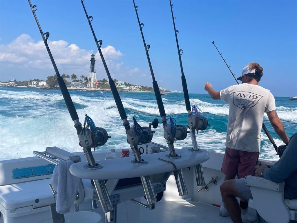 A person stands on a boat holding a fishing rod, perfecting the art of Hillsboro Inlet fishing. Four other rods are mounted nearby as the boat glides swiftly over the water. A distant lighthouse and palm trees provide a picturesque backdrop against a clear blue sky.