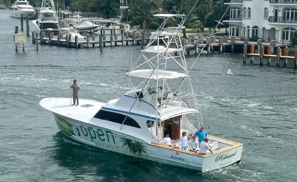 A fishing boat named "Rebound" sails along a waterway near Pompano Beach, featuring a tall tower and tropical-themed graphics. Several people are aboard, with one person standing at the bow. Luxurious waterfront homes provide a stunning backdrop, perfect for a memorable fishing charter experience.