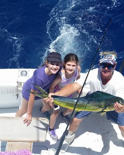 Two girls and a man on a boat hold up a freshly caught fish, smiling at the camera. With the ocean stretching behind them, fishing gear is scattered around—a classic day of Pompano drift fishing.