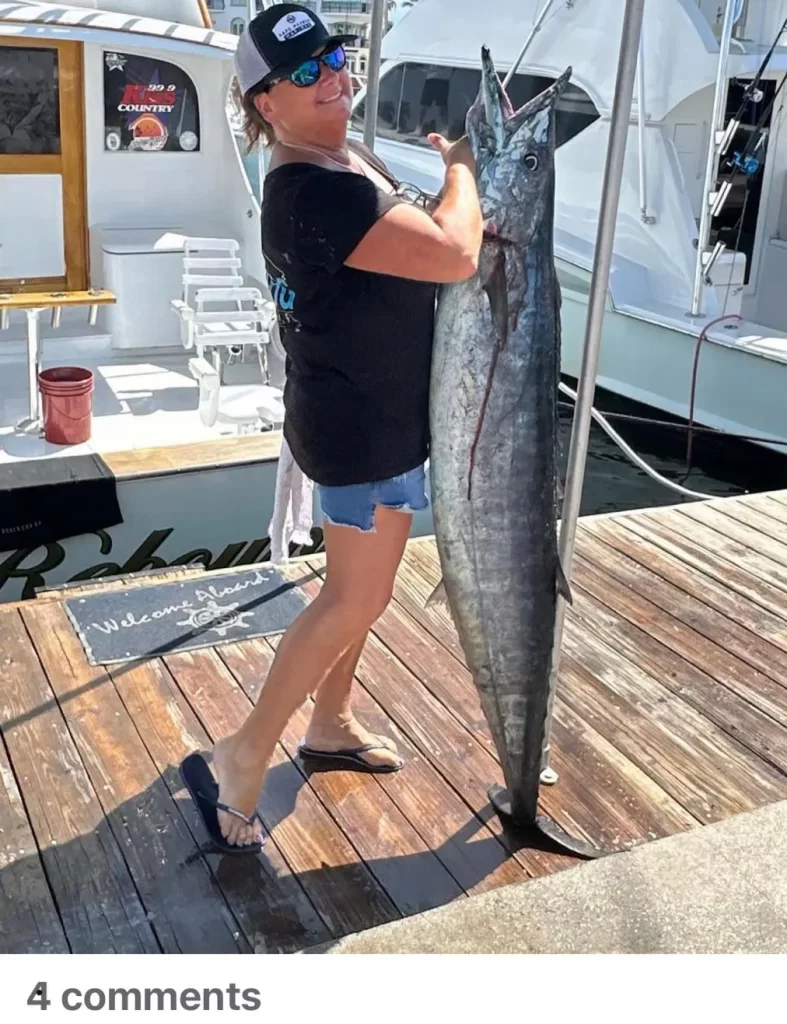 A person in casual clothing and a cap stands on a dock, proudly displaying an impressively large fish caught during a Hillsboro Inlet fishing adventure. Boats are moored in the background, and there's a small chalkboard sign promoting fishing charters Pompano Beach. The scene is bright and sunny.