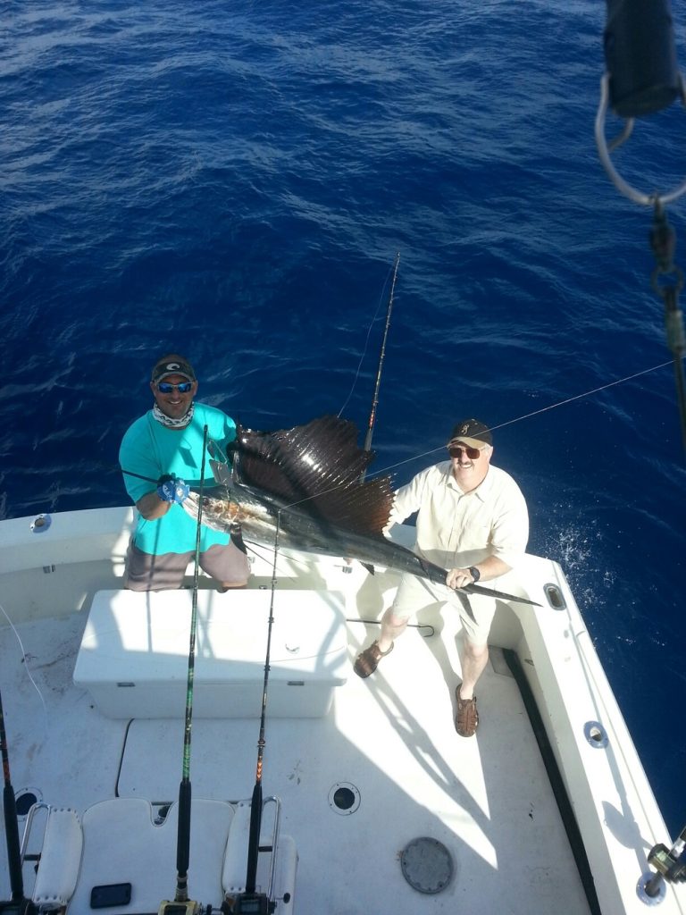 Two men stand on a boat off Hillsboro Inlet, proudly holding a large sailfish they caught. One wears a turquoise shirt, the other a white shirt and hat. Fishing rods line the boat, hinting at their successful Pompano Beach fishing charter adventure against the backdrop of the ocean.