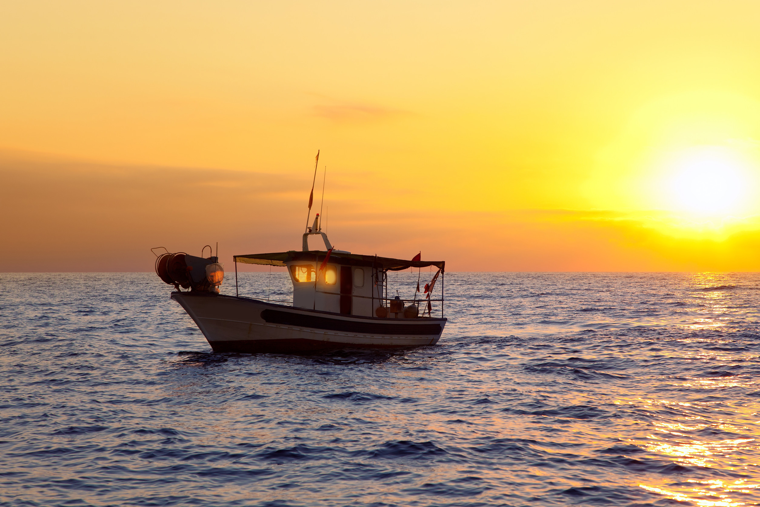 A fishing boat silhouetted against a vibrant sunset on the open sea. The sky is painted in warm hues of orange and yellow, while the calm waters reflect the colorful sky.