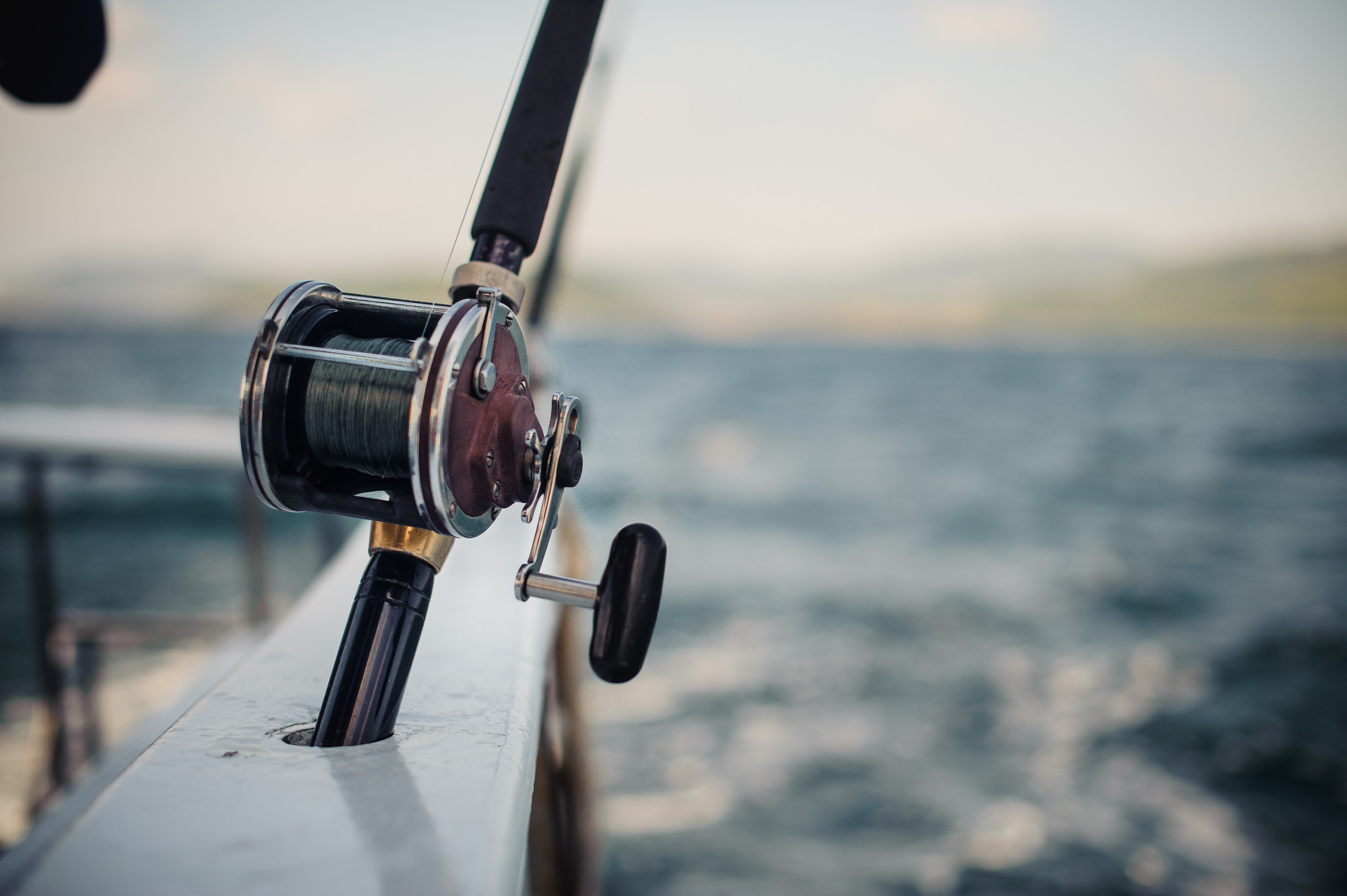 Close-up of a fishing reel mounted on the side of a boat, with a blurred view of the ocean and distant hills in the background. The scene conveys a sense of open water adventure and tranquility.