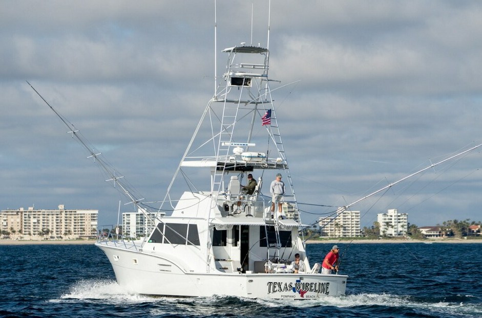 A white fishing boat named "Texas Shoreline" is cruising on the ocean. Several people are on board, with fishing rods extended. In the background, there are beachside buildings and a partly cloudy sky. An American flag flutters from the boat.