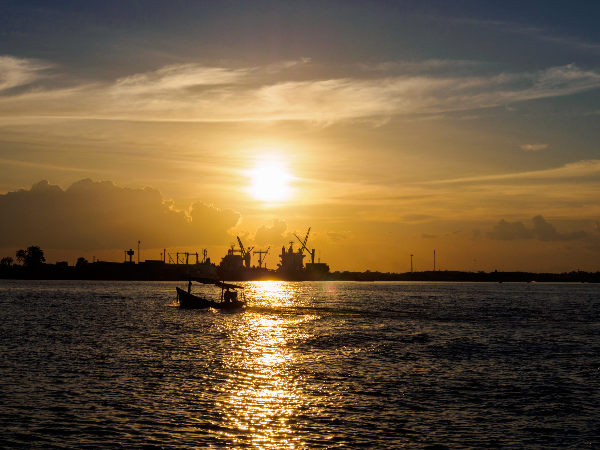 A small boat sails on a shimmering body of water at sunset, with golden sunlight reflecting on the water. Silhouettes of cranes and ships are visible on the horizon under a partly cloudy sky.