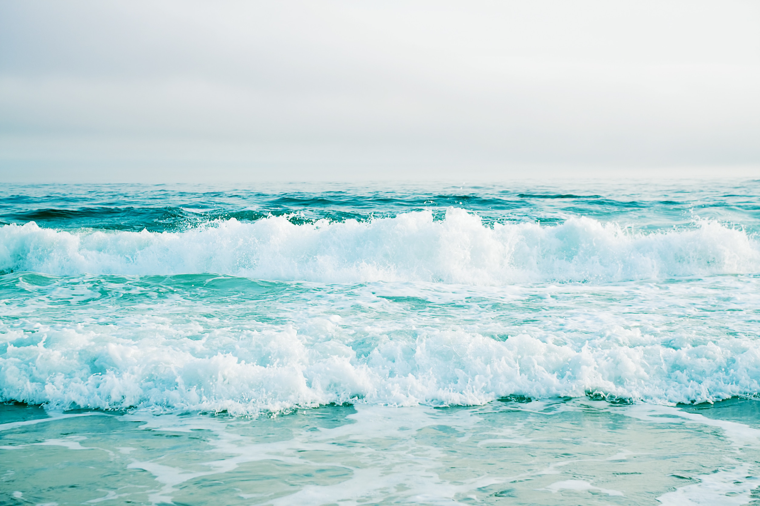 Ocean waves crashing onto a sandy shore under a cloudy sky. The water is a vibrant turquoise, and the waves create a frothy white foam as they hit the beach, capturing a serene and refreshing coastal scene.