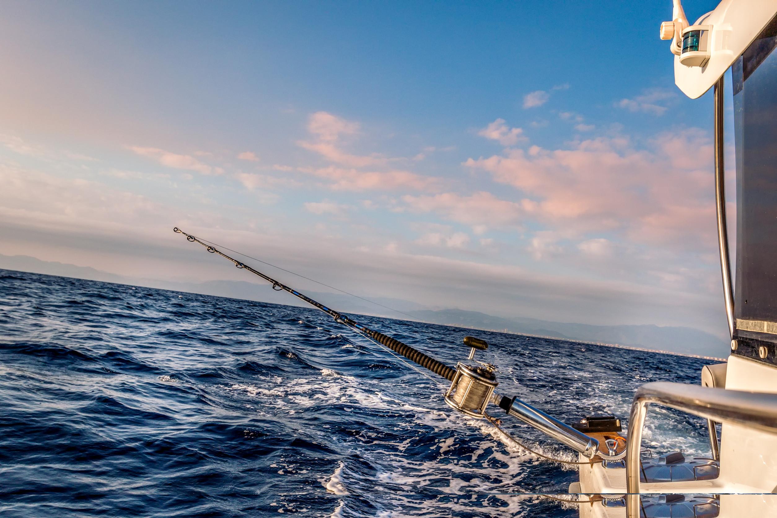 Fishing rod mounted on the side of a boat at sea during sunset. The sky is partly cloudy with a warm glow, and the ocean appears slightly choppy. Part of the boat is visible on the right side of the image.