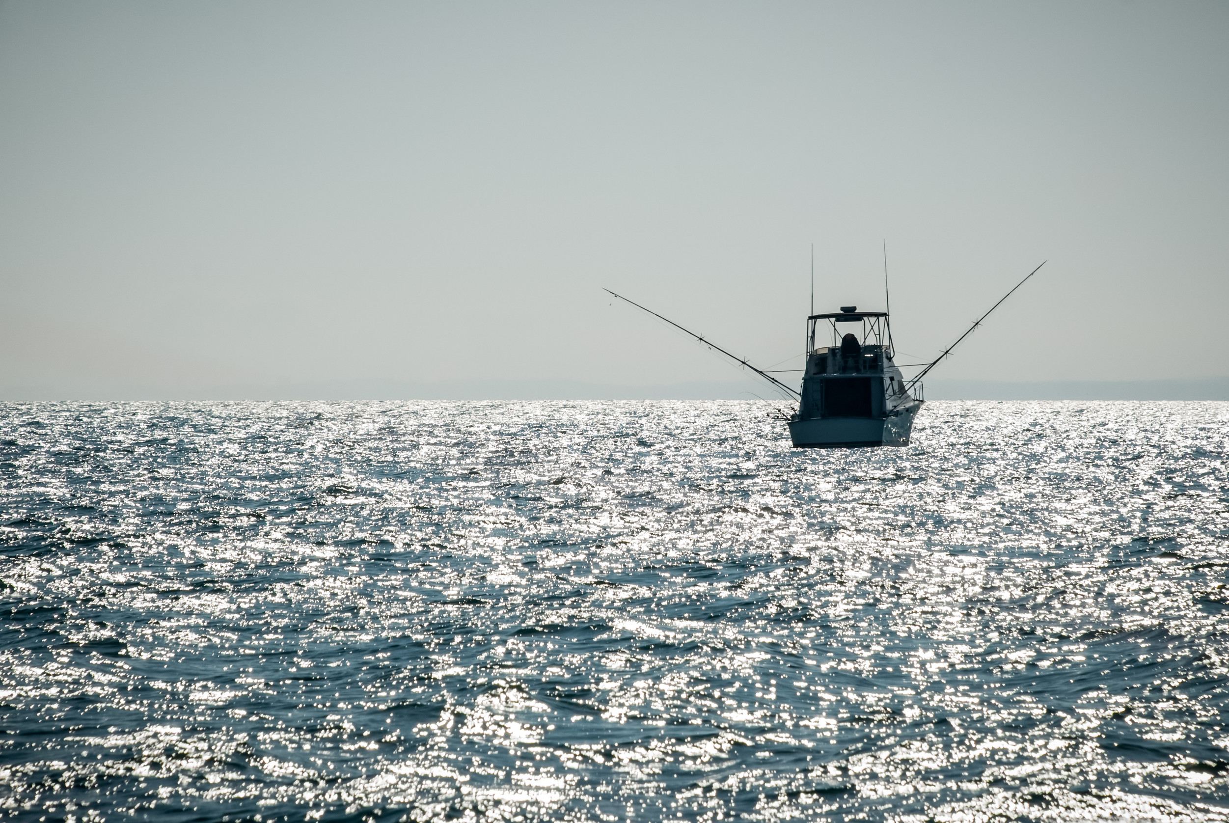 A boat with raised antennas is silhouetted against a shimmering, sunlit ocean. The horizon is faintly visible in the distance under a clear sky.