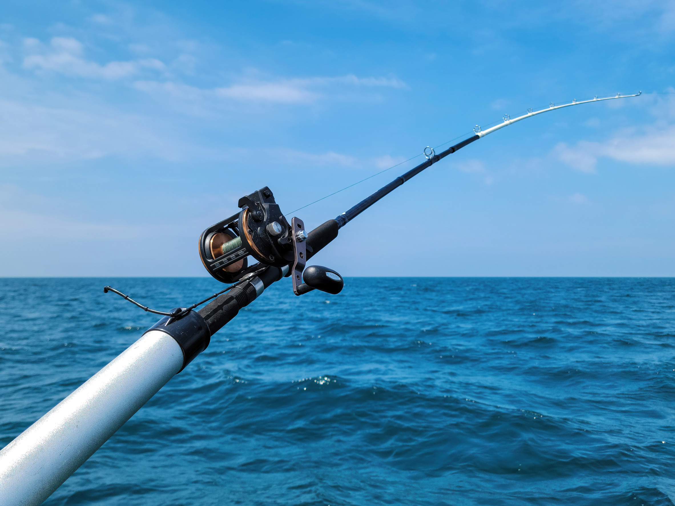 A fishing rod with a reel is extended over the side of a boat, bending slightly, with the blue ocean and a clear sky in the background.