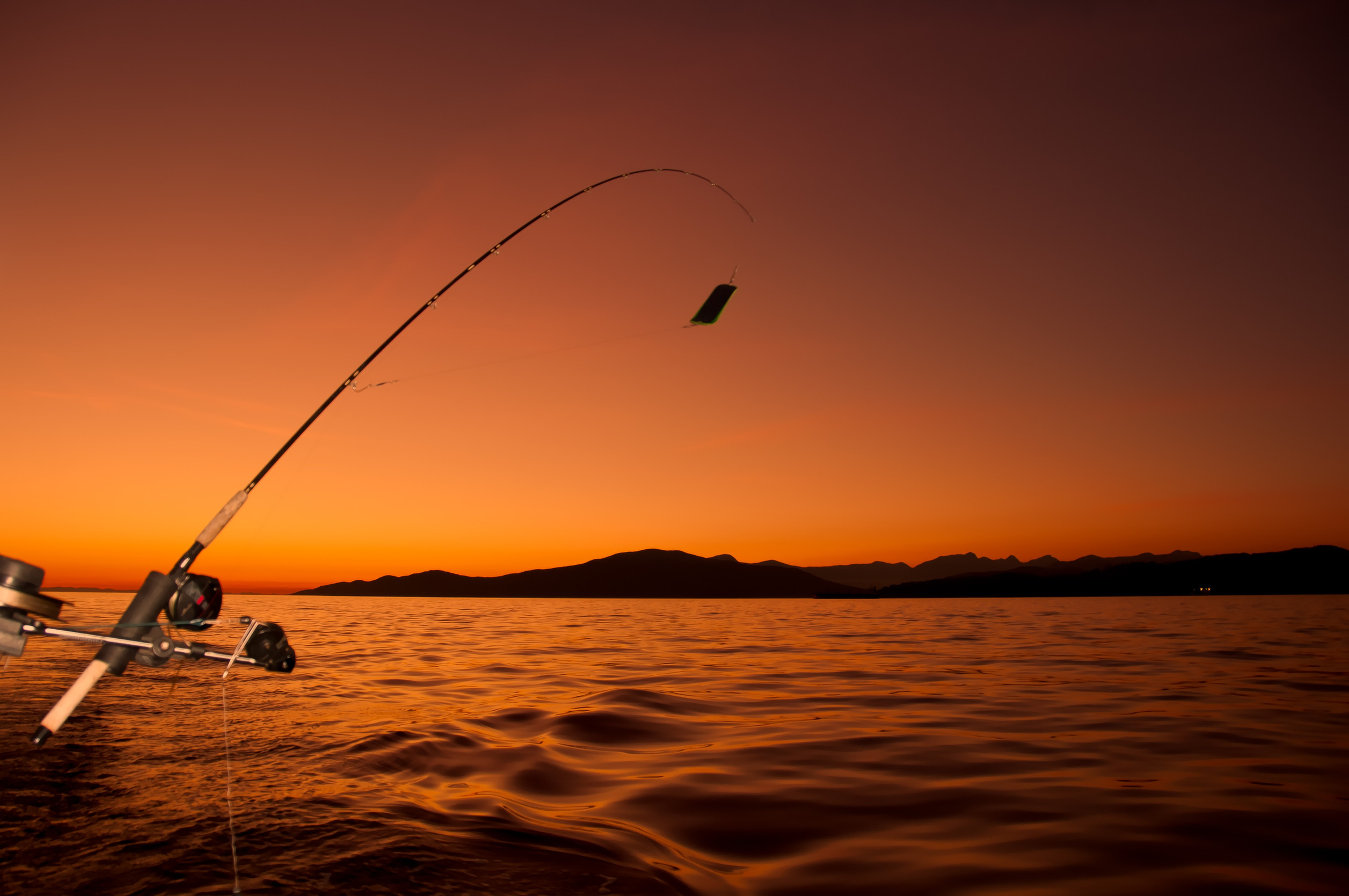 A fishing rod extended over calm water at sunset, with an orange sky and silhouetted mountains in the distance.