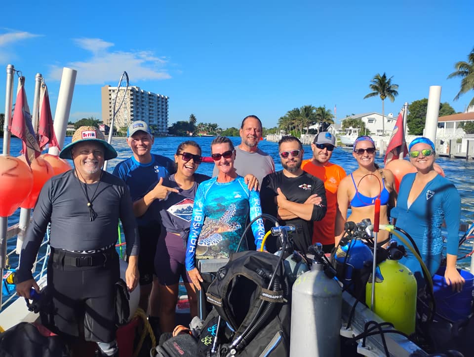 A group of nine smiling people in swim and dive gear pose on a boat with scuba tanks and diving equipment. The boat is on the water near palm trees and waterside buildings, under a clear blue sky.