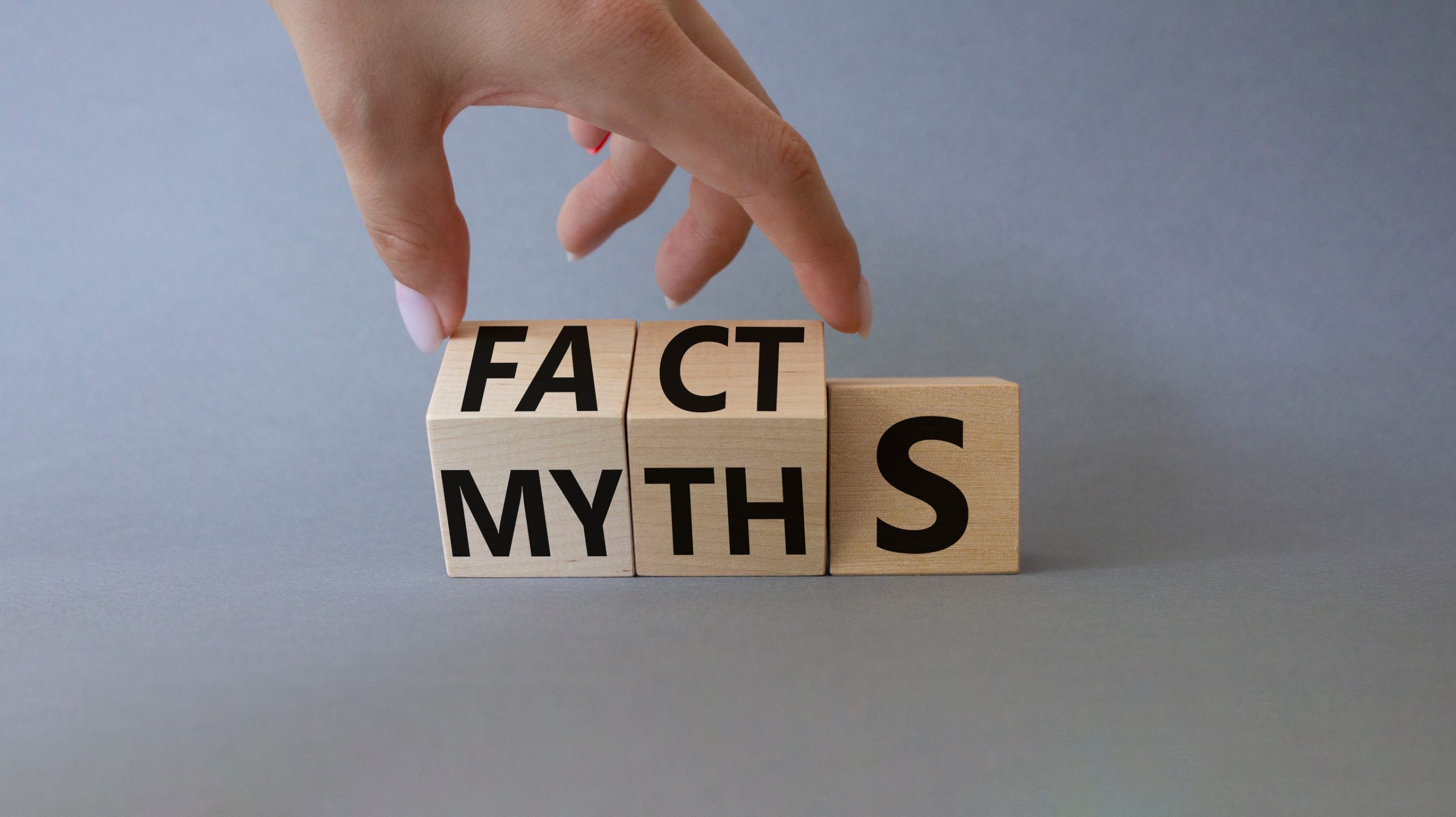 A hand rearranges wooden blocks that spell "MYTHS" and "FACTS" against a gray background, symbolizing the transition from myths to facts or vice versa.