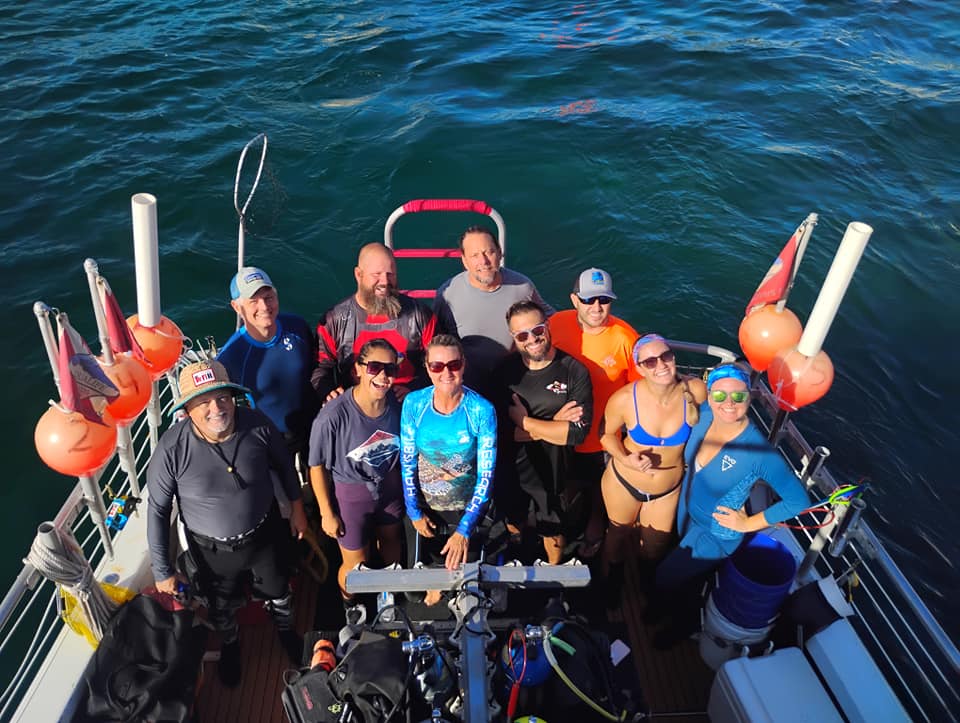 A group of eleven people in swimwear and diving gear smile on a boat surrounded by blue water, with orange buoys and dive equipment visible around them. The photo is taken from above.