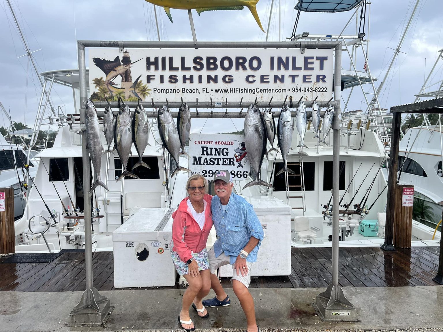 Two people pose and smile under a "Hillsboro Inlet Fishing Center" sign with a display of large fish hanging behind them at a marina, with fishing boats visible in the background on a cloudy day.