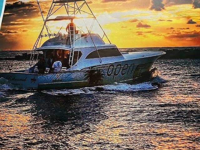 A white fishing boat with people onboard cruises through the water at sunset, with vibrant orange and yellow skies reflecting on the waves.
